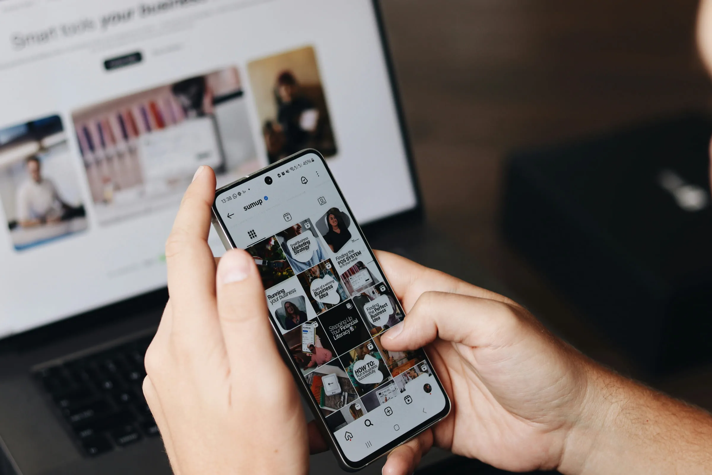 Zwei Hände halten ein Smartphone mit einem Social-Media-Feed, daneben ein aufgeklappter Laptop.